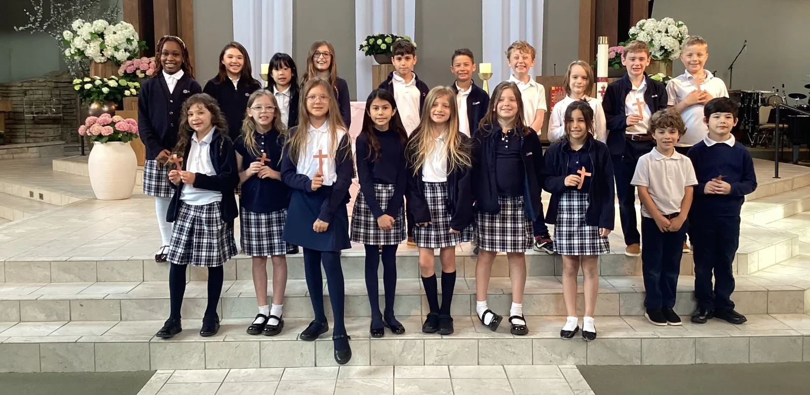 Holy Family Catholic Academy students in their school uniforms stand together on the altar steps in the church, smiling and holding small wooden crosses.